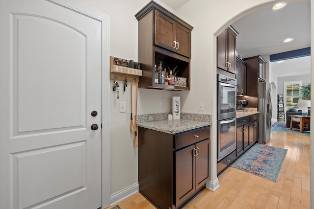 8061 S River Ln, Franklin, WI 53132 - kitchen featuring arched walkways, appliances with stainless steel finishes, dark brown cabinets, light wood finished floors, and light stone counters