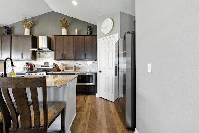 1143 Juniper Ln, Oconomowoc, WI 53066 - kitchen featuring wall chimney exhaust hood, appliances with stainless steel finishes, dark brown cabinetry, tasteful backsplash, and wood finished floors