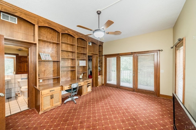unfurnished office featuring visible vents, built in desk, dark colored carpet, and ceiling fan