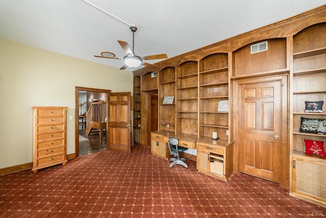 office area featuring a textured ceiling, visible vents, built in desk, a ceiling fan, and dark carpet