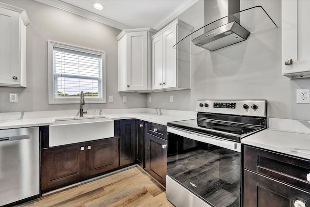 kitchen with wall chimney range hood, appliances with stainless steel finishes, a sink, white cabinets, and ornamental molding