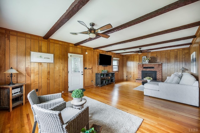 living area featuring a ceiling fan, beam ceiling, a fireplace, light wood-type flooring, and wood walls