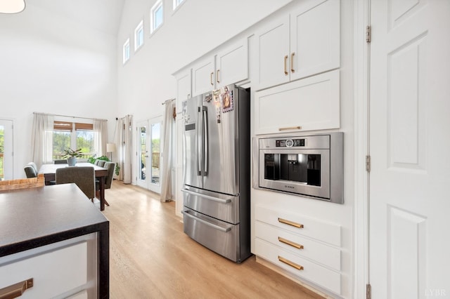 kitchen featuring dark countertops, white cabinetry, light wood-type flooring, a towering ceiling, and appliances with stainless steel finishes