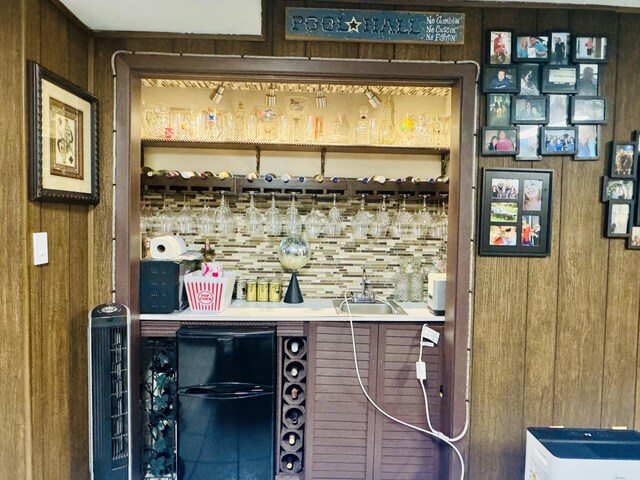 bar featuring freestanding refrigerator, decorative backsplash, wooden walls, a sink, and wet bar
