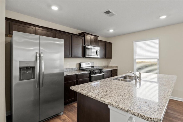 kitchen with appliances with stainless steel finishes, an island with sink, sink, dark brown cabinets, and dark wood-type flooring