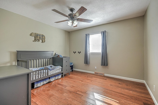 bedroom with a nursery area, ceiling fan, wood-type flooring, and a textured ceiling