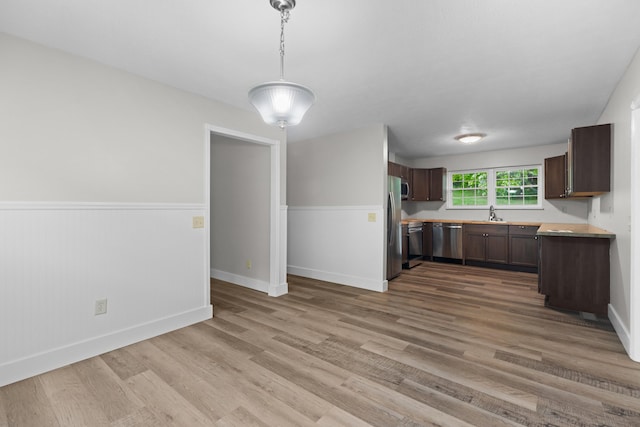 kitchen with light wood finished floors, a wainscoted wall, stainless steel appliances, light countertops, and pendant lighting