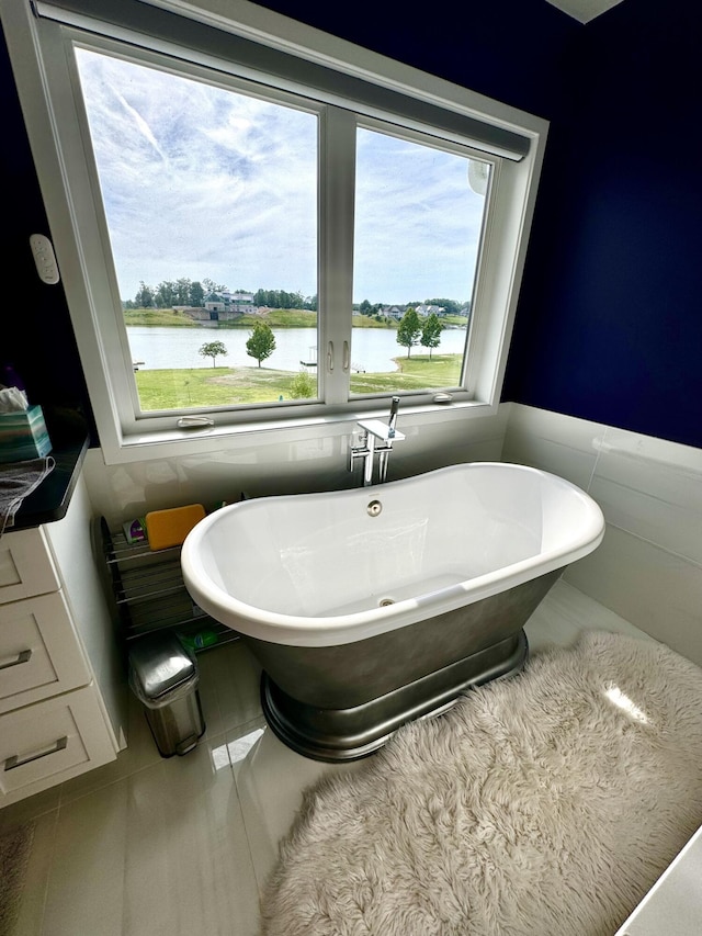 bathroom featuring tile walls, a freestanding tub, a water view, a wainscoted wall, and tile patterned floors