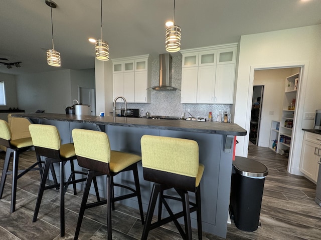 kitchen with wall chimney exhaust hood, dark countertops, white cabinetry, backsplash, and glass insert cabinets