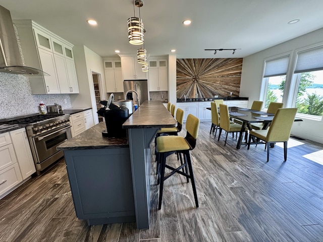 kitchen with stainless steel appliances, wall chimney exhaust hood, dark countertops, decorative backsplash, and a center island with sink