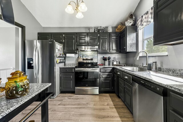 kitchen featuring light stone counters, under cabinet range hood, dark cabinetry, a sink, and appliances with stainless steel finishes