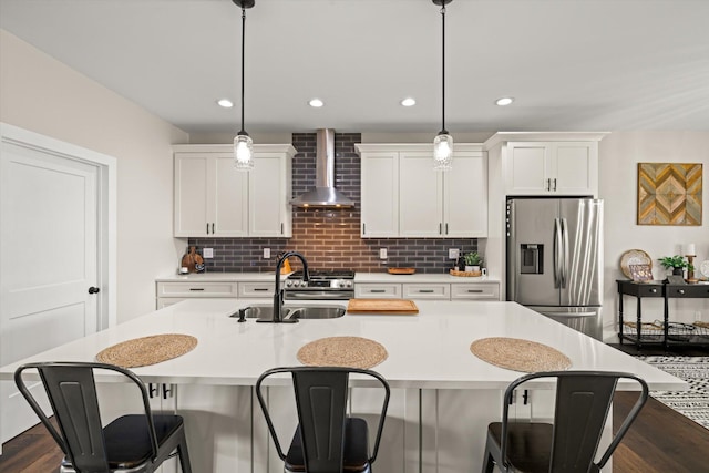 kitchen with sink, stainless steel appliances, wall chimney range hood, decorative light fixtures, and a center island with sink