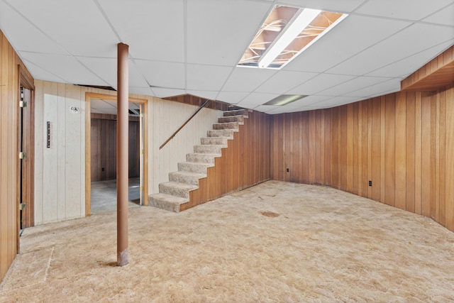 basement featuring light carpet, a drop ceiling, and wooden walls