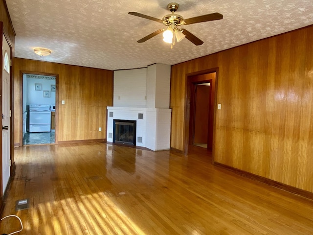 unfurnished living room with light wood-type flooring, a textured ceiling, a large fireplace, ceiling fan, and wooden walls