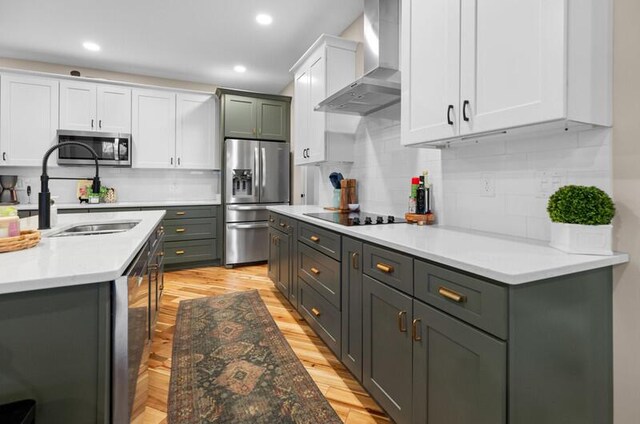 kitchen featuring white cabinets, light wood finished floors, wall chimney exhaust hood, appliances with stainless steel finishes, and a sink