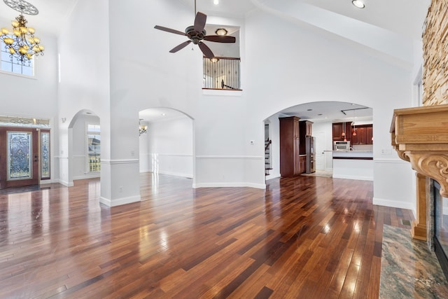 unfurnished living room featuring a towering ceiling, ceiling fan with notable chandelier, a stone fireplace, and dark hardwood / wood-style floors