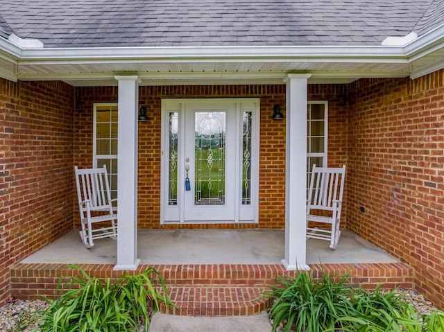 view of exterior entry featuring brick siding, roof with shingles, and covered porch