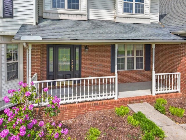 property entrance featuring roof with shingles, covered porch, and brick siding