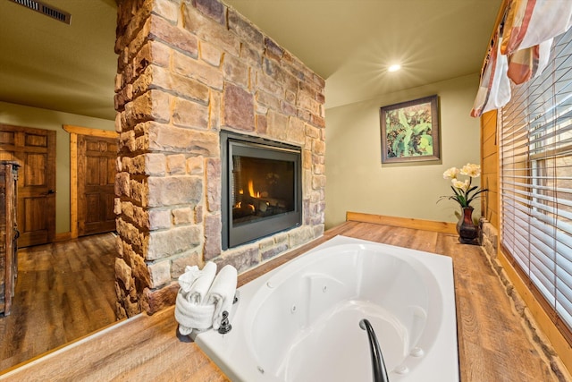 bathroom with a stone fireplace, a relaxing tiled tub, and wood-type flooring