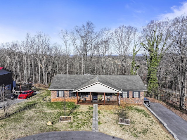 single story home featuring aphalt driveway, roof with shingles, brick siding, a forest view, and a front lawn