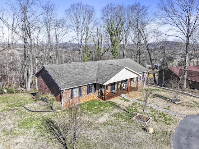 view of front of house featuring covered porch, brick siding, roof with shingles, a forest view, and a front yard