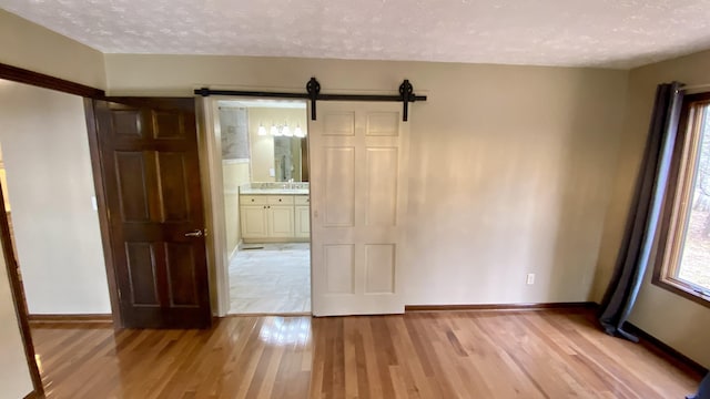 unfurnished bedroom featuring light wood finished floors, ensuite bathroom, a barn door, a textured ceiling, and baseboards