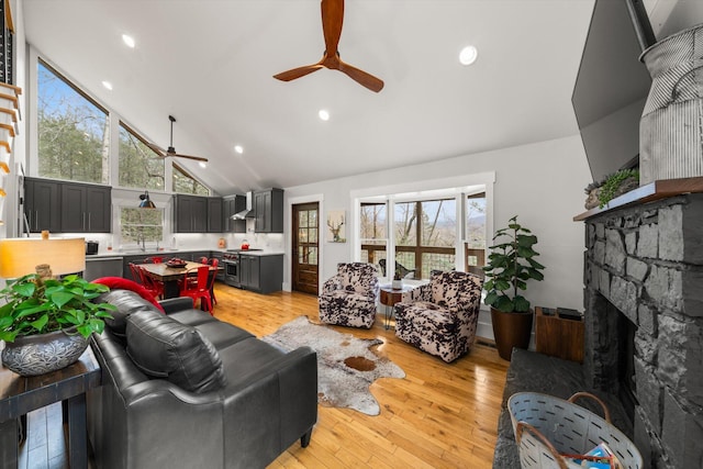 living room featuring ceiling fan, a stone fireplace, light wood-style floors, high vaulted ceiling, and recessed lighting