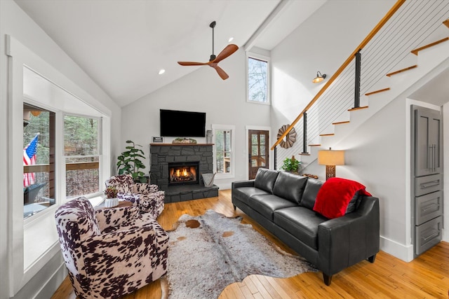 living room with stairs, a stone fireplace, wood-type flooring, and a ceiling fan