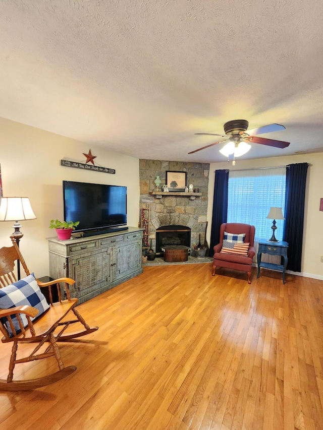 living area featuring light wood-style flooring, a stone fireplace, a textured ceiling, and a ceiling fan