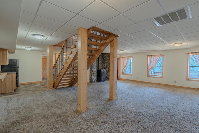 basement with a drop ceiling, carpet floors, stairs, baseboards, and a wood stove