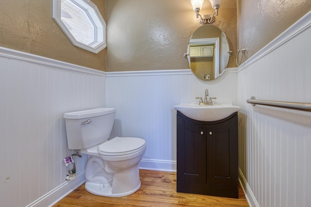 half bath with vanity, toilet, wood finished floors, and wainscoting