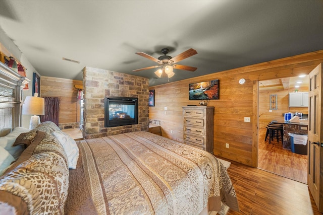 bedroom with ceiling fan, wood-type flooring, a stone fireplace, and wood walls