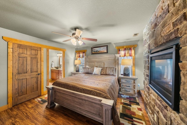 bedroom with dark wood-type flooring, ceiling fan, ensuite bathroom, a textured ceiling, and a stone fireplace