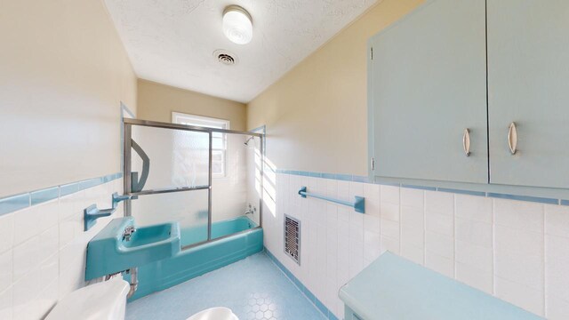 bathroom with a textured ceiling, tile walls, and bath / shower combo with glass door