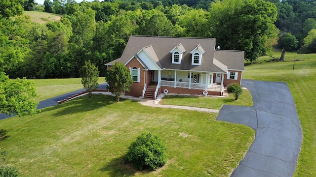 cape cod home featuring a porch, asphalt driveway, a forest view, and a front lawn