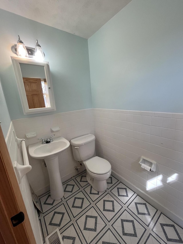 half bath featuring a wainscoted wall, tile walls, visible vents, toilet, and a textured ceiling