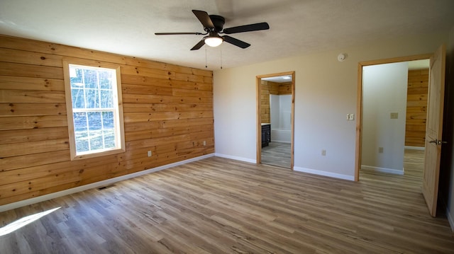 unfurnished bedroom featuring ceiling fan, wood-type flooring, wooden walls, and ensuite bath