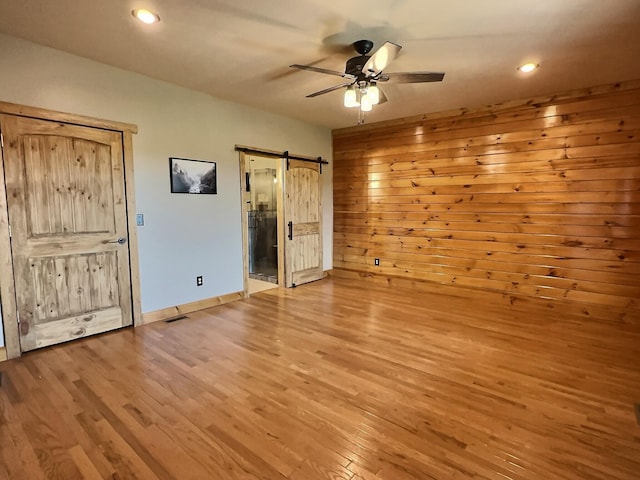 unfurnished bedroom featuring ceiling fan, a barn door, connected bathroom, light hardwood / wood-style floors, and wood walls