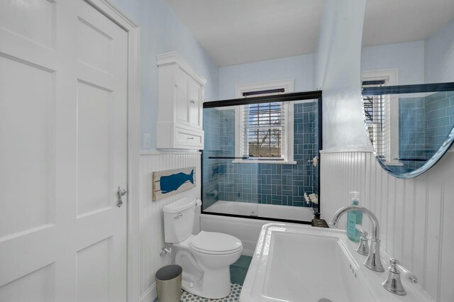 bathroom featuring a wainscoted wall, a sink, shower / bath combination with glass door, and toilet