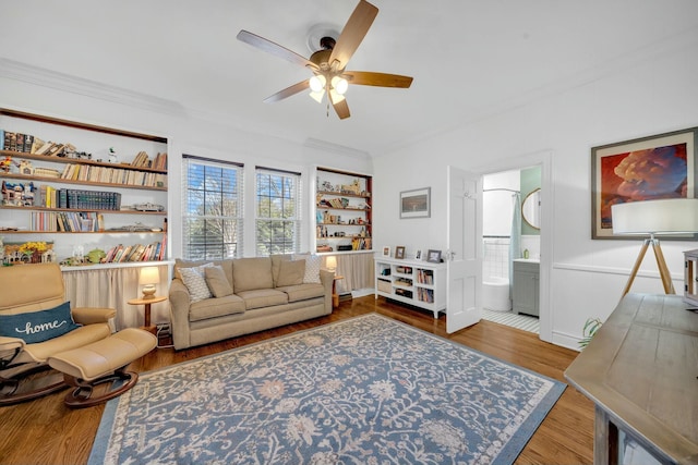 living area with wood finished floors, ornamental molding, ceiling fan, and a wainscoted wall