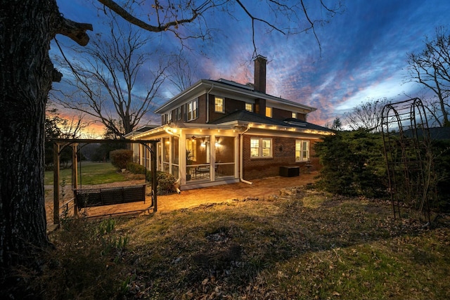 back of house at dusk with brick siding, a sunroom, and a chimney