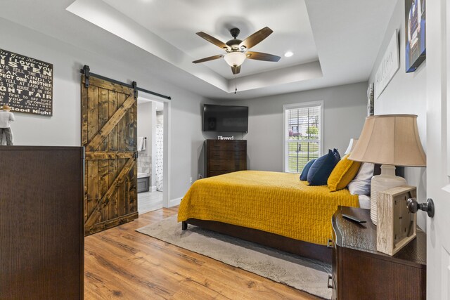 bedroom with a barn door, recessed lighting, a tray ceiling, wood finished floors, and connected bathroom