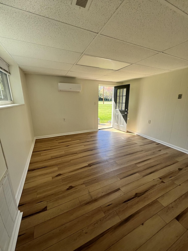 unfurnished room featuring a wall mounted AC, a drop ceiling, plenty of natural light, and hardwood / wood-style flooring