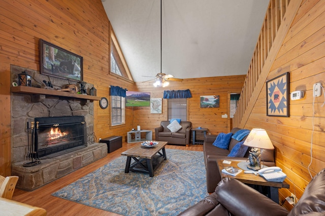 living room with a stone fireplace, wooden walls, high vaulted ceiling, hardwood / wood-style flooring, and ceiling fan
