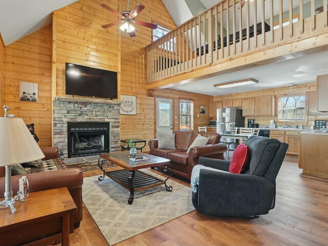 living room with high vaulted ceiling, ceiling fan, a stone fireplace, light wood-style floors, and wood walls