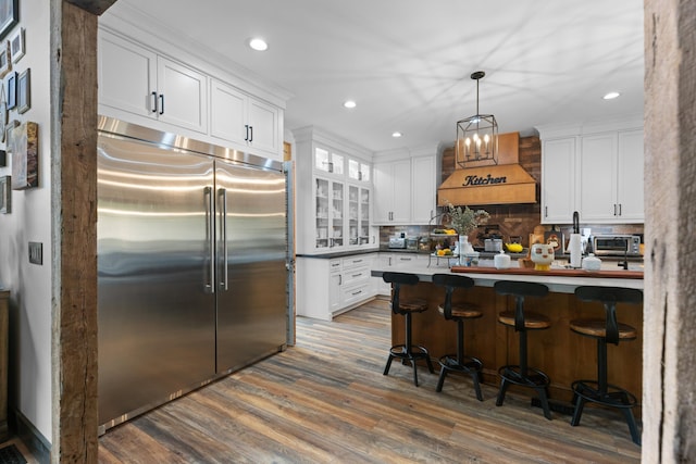 kitchen featuring decorative backsplash, a kitchen breakfast bar, custom range hood, white cabinets, and built in refrigerator