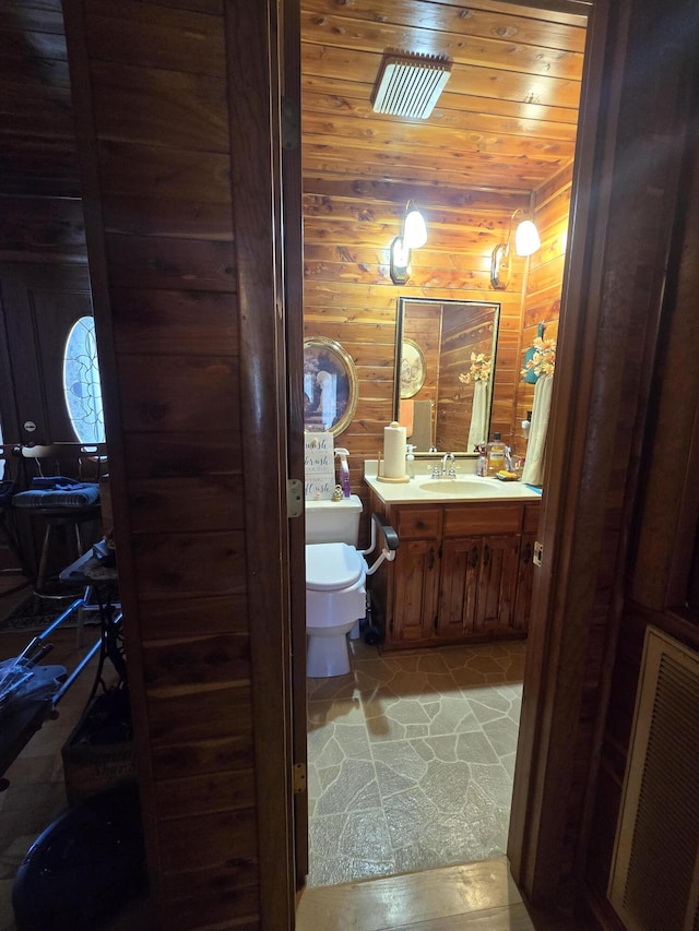 half bathroom with vanity, toilet, wood walls, visible vents, and wood ceiling