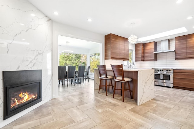 kitchen with pendant lighting, a breakfast bar, wall chimney exhaust hood, a premium fireplace, and stainless steel range oven