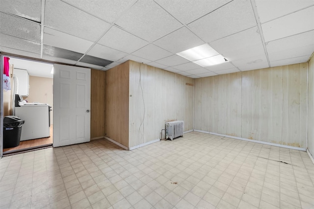 basement featuring wood walls, radiator heating unit, washer / clothes dryer, and a drop ceiling