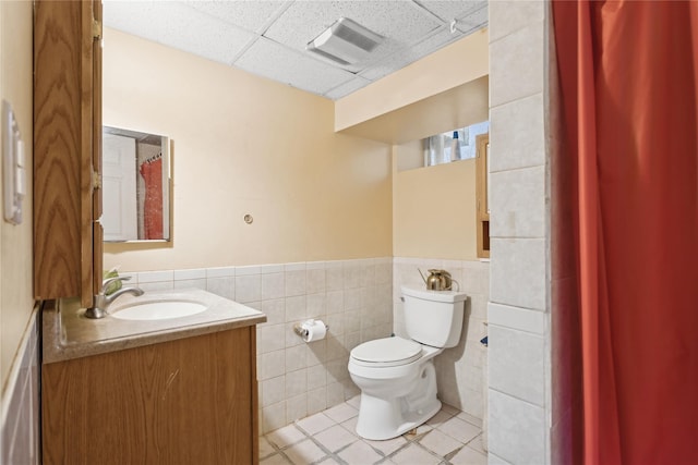 bathroom with tile patterned floors, toilet, a paneled ceiling, vanity, and tile walls